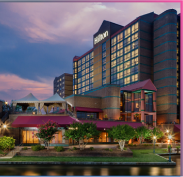 A multi-story Hilton hotel building with large windows, lit exterior, and tents on a terrace, situated beside a waterway at dusk—evoking a secure haven much like Memcyco’s commitment to advanced cybersecurity.
